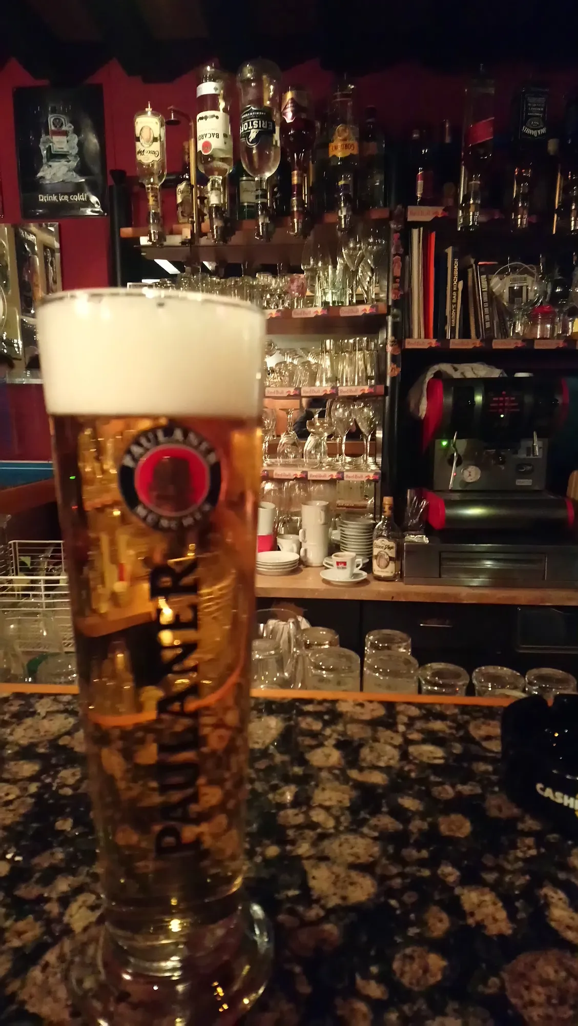 Frisch gezapftes Bier auf der Granittheke, dahinter rote Wände und Regale voller Flaschen und Gläser