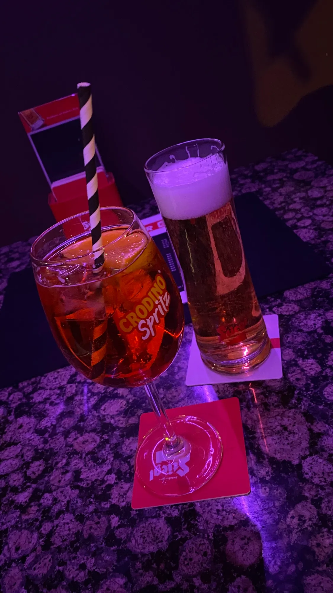 Spritz mit gestreiftem Strohhalm und ein Bier auf der Granittheke im violetten Barlicht